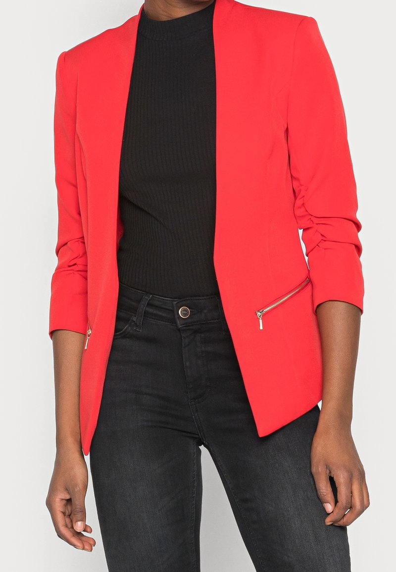 Blazer rojo de manga larga, con un corte ajustado, dos bolsillos laterales con cremallera y una textura lisa, combinado con una camiseta negra acanalada y vaqueros oscuros.