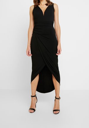 Sleeveless black midi dress with V-neck, ruched waist, tulip hem, worn with black ankle-strap high-heeled sandals, standing on white floor.