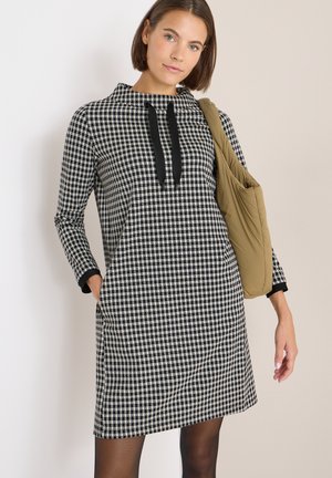 Schwarz-weißer karierten Kleid mit Trichterkragen, langen Ärmeln, Seitentaschen und kontrastierenden schwarzen Manschetten, kombiniert mit einer braunen Schultertasche.