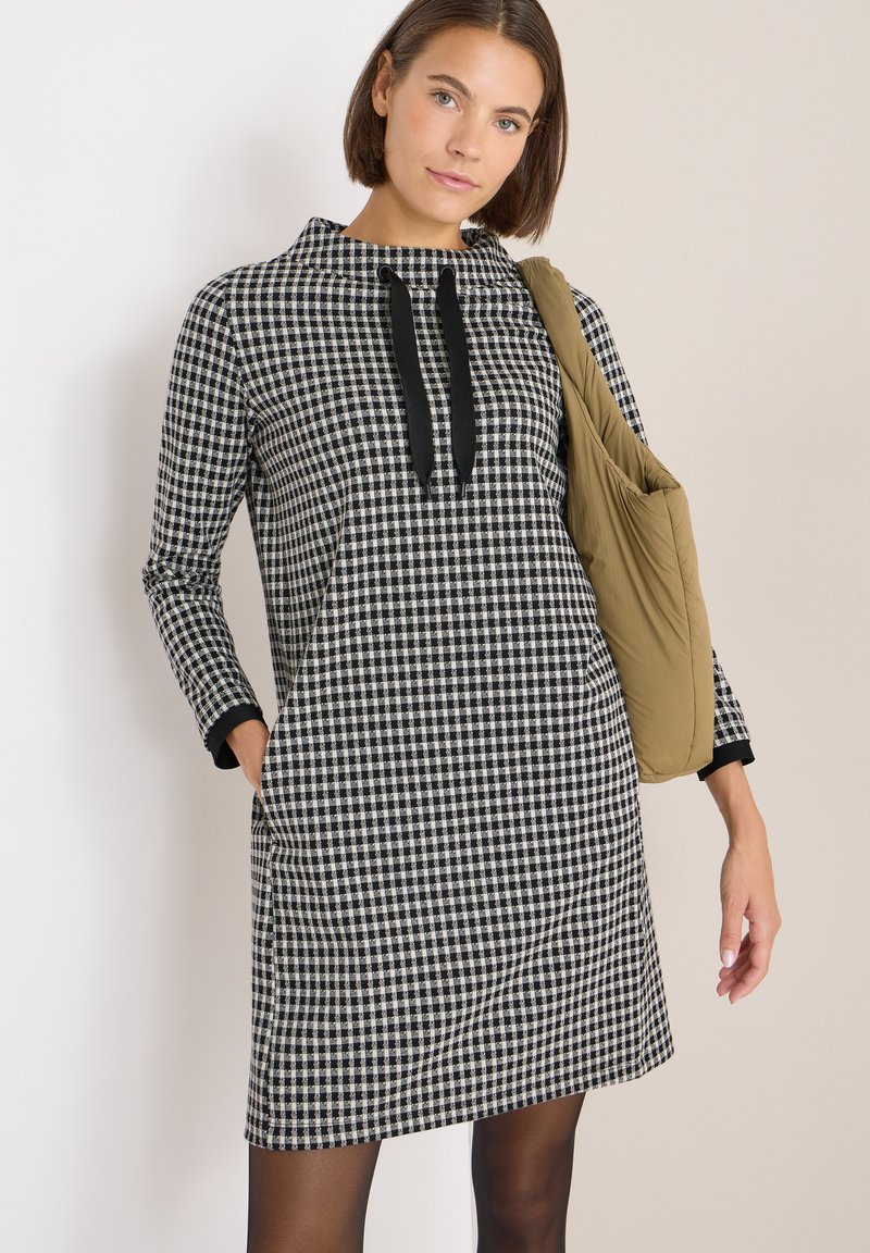 Czarna i biała sukienka w kratę z kołnierzem w kształcie lejka, długimi rękawami, bocznymi kieszeniami i kontrastującymi czarnymi mankietami, zestawiona z brązową torebką na ramię.