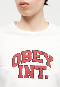 Κοντινό πλάνο ενός ατόμου που φοράει λευκό πουκάμισο με μεγάλο κόκκινο έντονο κείμενο "OBEY INT." στο στήθος.