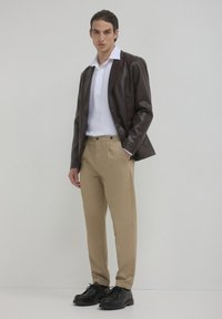 Brauner Kunstleder-Blazer über einem weißen Polo-Shirt, kombiniert mit hellbeigen Hosen und schwarzen Schnürschuhen. Glatte Texturen, taillierte Passform.