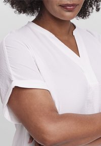 Weißes kurzärmliges Blusenshirt mit einem V-Ausschnitt und umgeschlagenen Ärmeln. Hergestellt aus leichtem Stoff mit einer subtilen Textur.