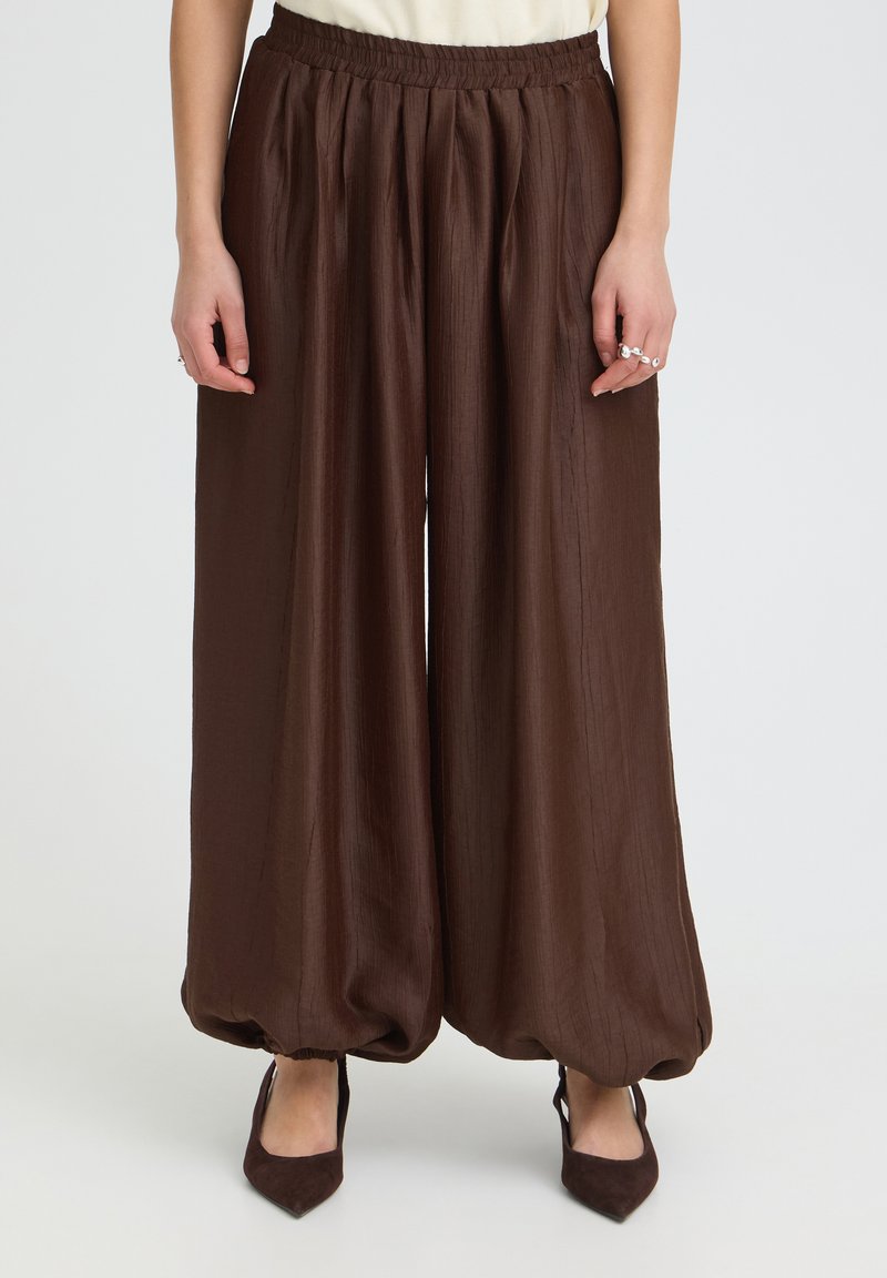 Pantalon marron ample avec poignets élastiques froncés porté avec des chaussures pointues marron foncé et un haut blanc, montré de la taille aux pieds.