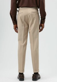 Beige op maat gemaakte pantalon met rechte pijpen, voorzien van twee achterzakken met knoopsluitingen en opgerolde zomen, gecombineerd met donkere schoenen.