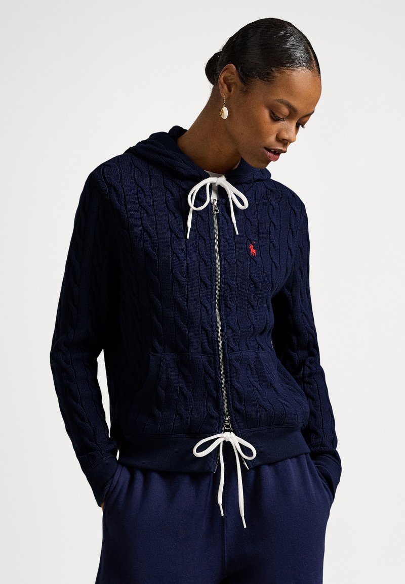 Ženska v mornarsko modri pleteni jopa z zadrgo in belimi vrvicami ter ujemajočimi mornarsko modrimi hlačami, pogled usmerjen navzdol z rokami v žepih.