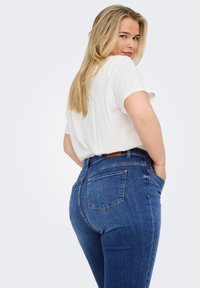 Blue high-waisted jeans with a textured denim finish, back pockets, and a brown leather patch at the waistband, paired with a white t-shirt.