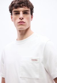 Νεαρός άνδρας με κοντά σγουρά μαλλιά που φοράει ένα λευκό t-shirt Calvin Klein με τσέπη στο στήθος και λογότυπο, σε απλό λευκό φόντο.