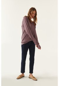 Maglietta lunga mauve di taglio morbido, jeans slim blu scuro e mocassini beige con dettagli in metallo, su uno sfondo chiaro.