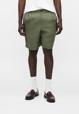 Άνδρας που φοράει χακί πλισέ σορτς, λευκές κάλτσες μέχρι τη μέση της γάμπας, καφέ δερμάτινα μοκασίνια και λευκό πουκάμισο, στέκεται μπροστά σε απλό φόντο.