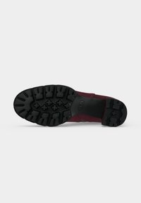 Semelle de chaussure avec des motifs de semelle noirs profonds et une tige bordeaux visible de côté, présentée sur un fond clair uni.