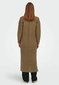 Robe tricotée longue de couleur vert olive avec une finition texturée, des manches côtelées et un ourlet. Portée avec un pantalon noir et des baskets basses, vue de dos.