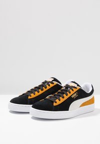 Svarta och orange mockaskor med vita detaljer, med en strukturerad sula och svarta snören. Logotypen visas på tungan.