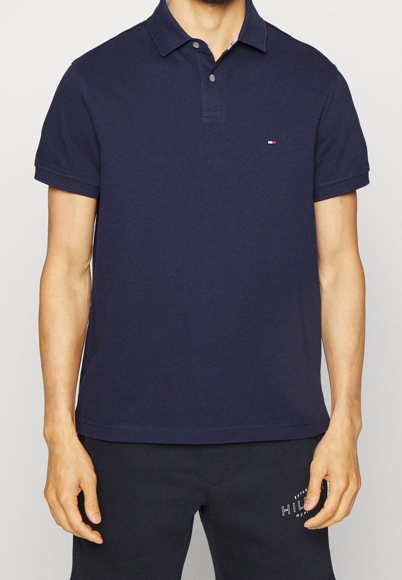 Homme portant un polo bleu marine à manches courtes et un pantalon noir avec un logo blanc sur la cuisse gauche.