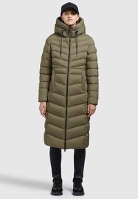 Kvinde iført lang olivengrøn quilted pufferjakke med hætte, sort kasket, sorte bukser og sorte sneakers står foran en grå baggrund.