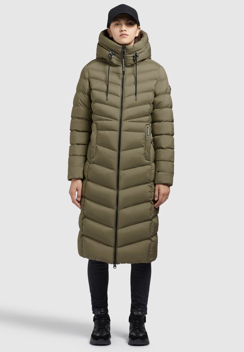 Kvinde iført lang olivengrøn quilted pufferjakke med hætte, sort kasket, sorte bukser og sorte sneakers står foran en grå baggrund.