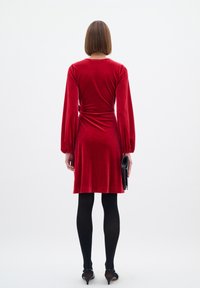 Röd sammetsklänning med långa puffärmar, figursydd midja och A-linjeformad silhuett. Modellen bär en svart clutch och har på sig svarta strumpbyxor och klackar.