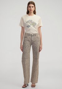 Camiseta de algodón beige con un estampado gráfico floral. Jeans de pierna ancha color marrón claro con un patrón moteado, combinados con sandalias negras.