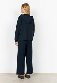 Dunkelblaues Kapuzenshirt mit Kapuze, lockere Passform, kombiniert mit weit geschnittenen Hosen. Weicher Stoff, schlichtes Design, flache Slipper mit Nieten.