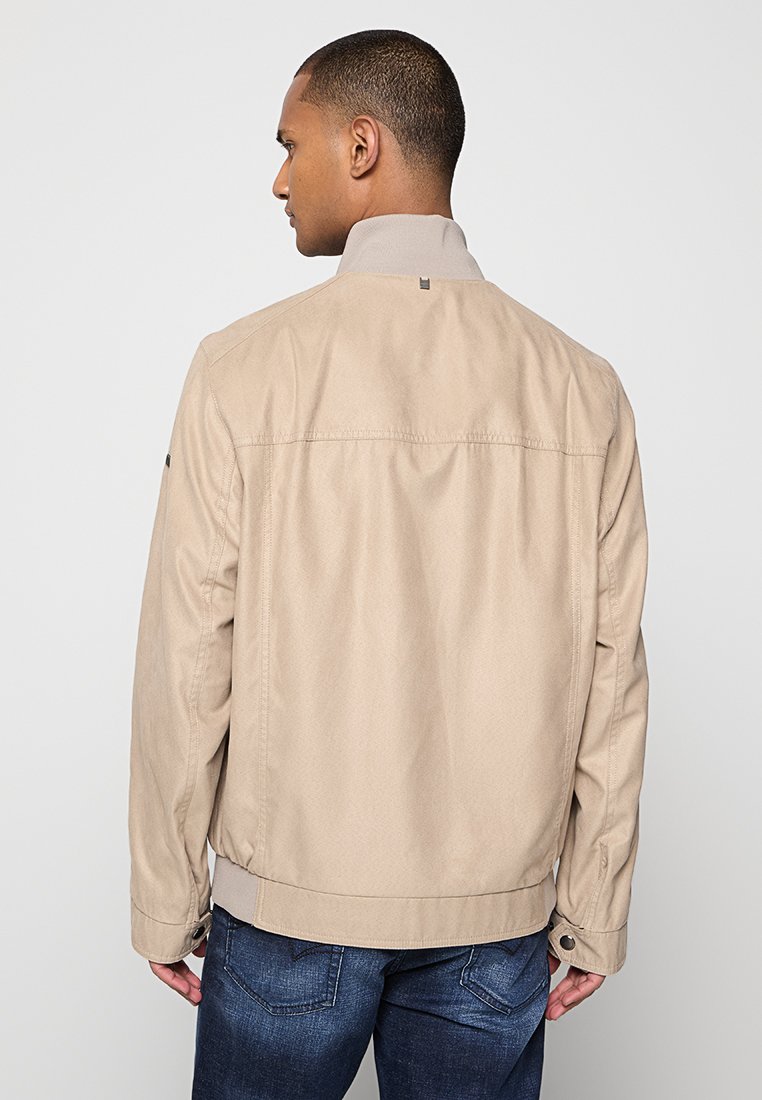 Veste légère beige avec un col haut, en tissu texturé et avec des poignets à boutons, vue de dos sur un fond neutre.