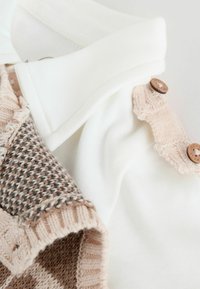 Kokvilnas un adīta apģērba gabals ar baltu apkakli, beiga pogām un rakstainu brūnu audumu ar faktūrām un neitrālām krāsām.