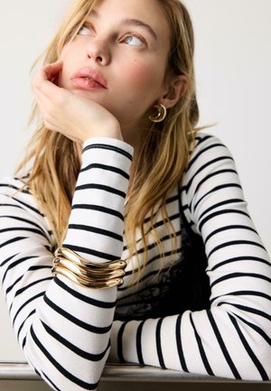 Jonge vrouw met blond haar, gekleed in een zwart-wit gestreept shirt met lange mouwen, een gouden armband en gouden halvemaanvormige oorbellen, leunt met haar gezicht op haar hand.