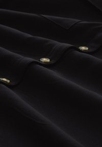 Black shirt fabric with smooth texture, featuring prominent gold buttons. The fabric drapes gently, showcasing sleek curves and hemline.