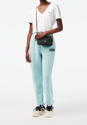 Wit V-hals T-shirt, lichtblauwe joggingbroek met zwarte letters langs de zijkant, en een zwarte crossbodytas. Afgewerkt met witte en navy sneakers.