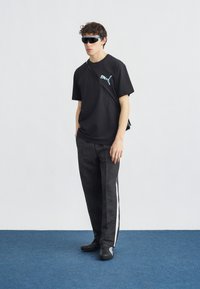 Νέος άνδρας με μαύρο μπλουζάκι Puma, μαύρο παντελόνι με λευκή ρίγα, μαύρα παπούτσια, τσάντα χιαστί και γυαλιά ηλίου με μπλε φακούς, στέκεται σε μπλε χαλί.