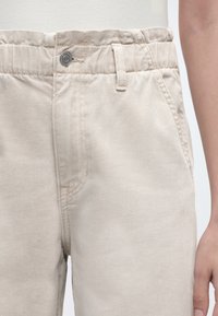 Κοντινό πλάνο μπεζ παντελονιού με ελαστική μέση, μπροστινό κουμπί, θηλιά για ζώνη, πλαϊνή τσέπη και εμφανές μέρος του χεριού ενός ατόμου.