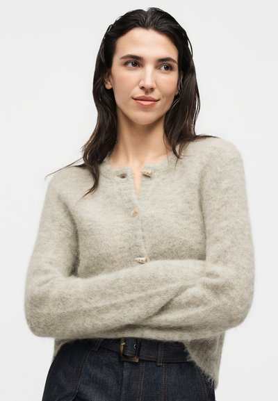 Femme aux longs cheveux foncés portant un cardigan beige duveteux avec des boutons en bois et un pantalon sombre taille haute, bras croisés, regardant sur le côté.