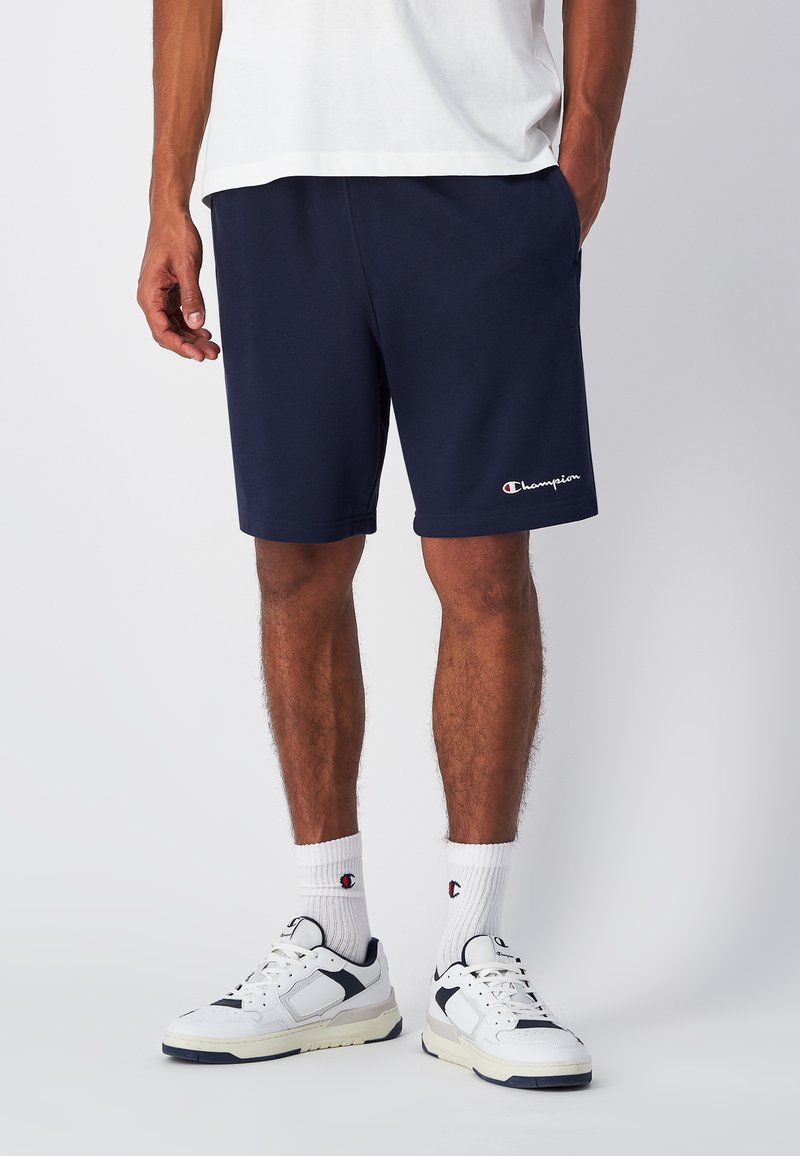 Marineblaue Shorts mit elastischem Bund, besticktem Logo am linken Bein, kombiniert mit weißen Turnschuhen und weißen Socken mit Logo-Akzenten.