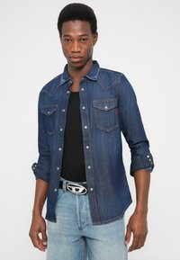 Chemise en denim foncé avec fermeture boutonnée sur le devant, deux poches poitrine et quincaillerie argentée. Associée à un T-shirt noir et un jean bleu clair.