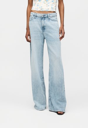 Osoba nosząca jasnoniebieskie, szerokie spodnie jeansowe z wysokim stanem i krótki kwiatowy top, stojąca z rękami swobodnie opuszczonymi wzdłuż ciała.