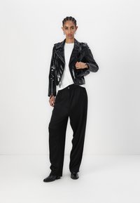 Veste de moto en cuir verni noir sur un t-shirt blanc, accompagnée de pantalons noirs amples et de bottes noires. Fond non réfléchissant.