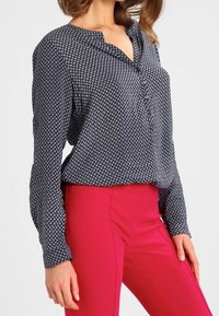 Blusa a maniche lunghe di colore blu navy con motivo, con un'unica chiusura a bottone sul davanti e una vestibilità comoda, abbinata a pantaloni aderenti di un brillante colore rosa.}}