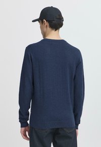 Maglione navy a girocollo con maniche lunghe, texture liscia, polsini e orlo a coste. Indossato con un berretto da baseball scuro.