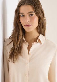 Jeune femme aux longs cheveux bruns portant une chemise beige clair à boutons, posant devant un fond clair.