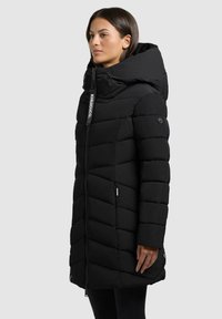 Svart dunjakke med høy krage og hette, med glidelås foran, skrå polstring og logodetaljer på glidelåsbåndet.