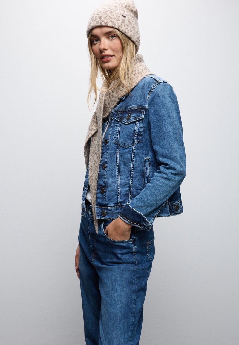 Giacca in denim di blu medio con due tasche al petto, chiusura con bottoni e cuciture a contrasto, abbinata a una sciarpa e un cappello con stampa leopardata beige chiaro.