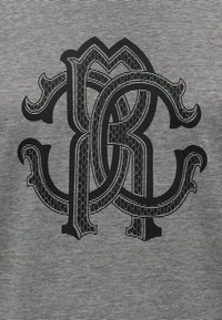 Πλέκονται περίτεχνα μαύρα γράμματα «Μ», «C» και «R» με μοτίβο διαμαντιού, τοποθετημένα πάνω σε λεπτό φόντο με μαύρες και λευκές ρίγες.