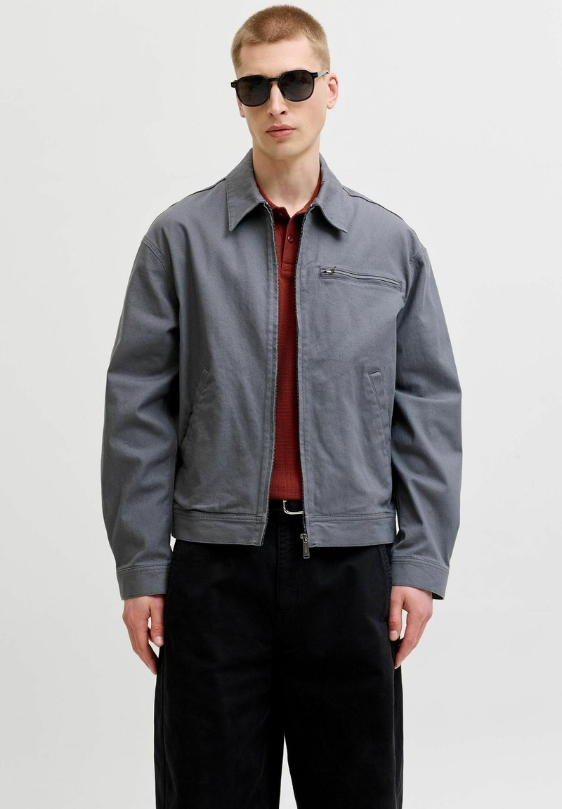 Jeune homme portant des lunettes de soleil noires, une veste grise zippée, une chemise rouge à col et un pantalon noir, debout devant un fond clair uni.