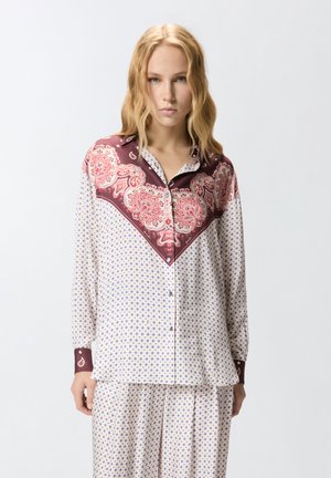 Femme blonde portant une chemise blanche à boutons et un pantalon assorti avec de petits motifs bleus et un imprimé cachemire bordeaux sur les épaules et les poignets.