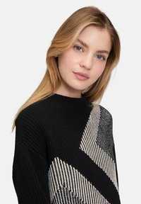 Schwarzer und cremefarbener Strickpullover mit einem geometrischen Zickzackmuster. Ribbeltextur und Rundhalsausschnitt, mit langen Ärmeln und lockerer Passform.
