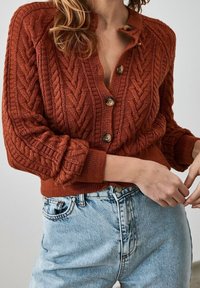 Rust-colored knitted cardigan with a cable pattern, featuring a ribbed hem and cuffs, and five buttons on the front. Worn with light blue jeans.