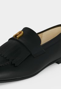 Mocassin en cuir noir avec détail à franges et boucle décorative dorée sur le dessus, présentant une surface lisse et polie ainsi qu'une doublure intérieure beige clair.