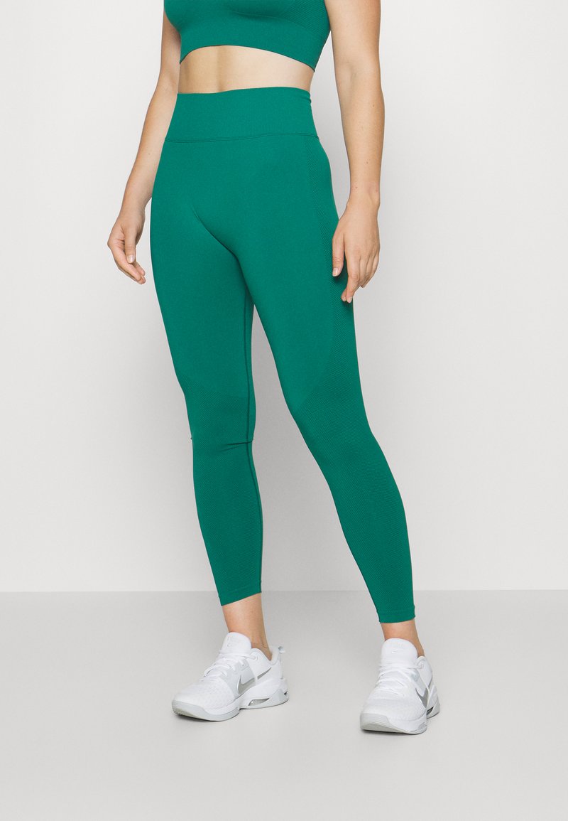 Grønne høytliv leggings med en glatt tekstur og en tettsittende design, kombinert med hvite joggesko med grå detaljer.