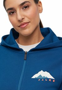 Felpa blu con zip e tasca a marsupio, caratterizzata da una grafica di montagne e la scritta "FALKE" in ricamo arancione sul lato sinistro del petto.