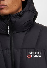 Doudoune noire avec un col montant, dotée d'une fermeture éclair sur le devant et du logo "SOUTH POLE" en blanc et rouge sur la poitrine. Surface texturée.