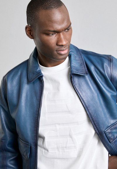 Veste en cuir bleue avec un col, fermeture éclair à l'avant et poches latérales, portée sur un T-shirt blanc avec un motif en relief subtil.